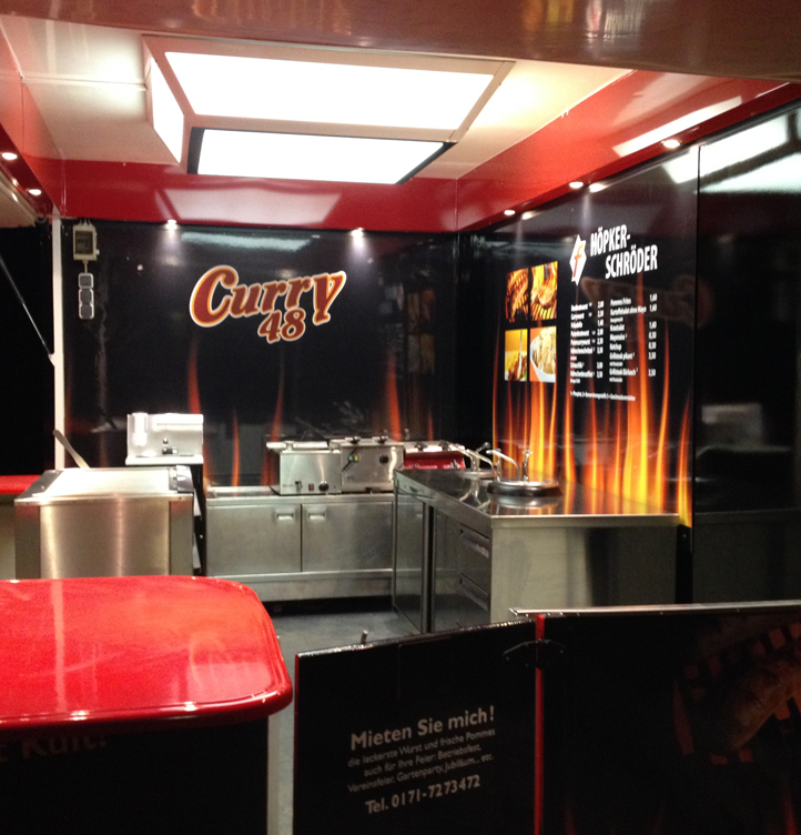 Imbissstand mit glänzenden Edelstahlflächen und rotem Tresen vor schwarzer Wand