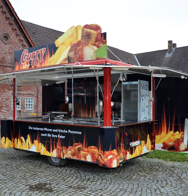 Ein Imbisswagen mit bunter Werbung für Currywurst und Pommes auf Kopfsteinpflaster